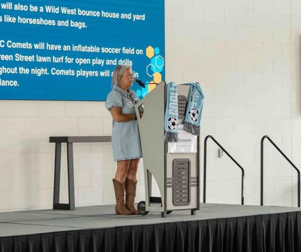 Person at a podium on a stage, speaking into a microphone beside a large screen.