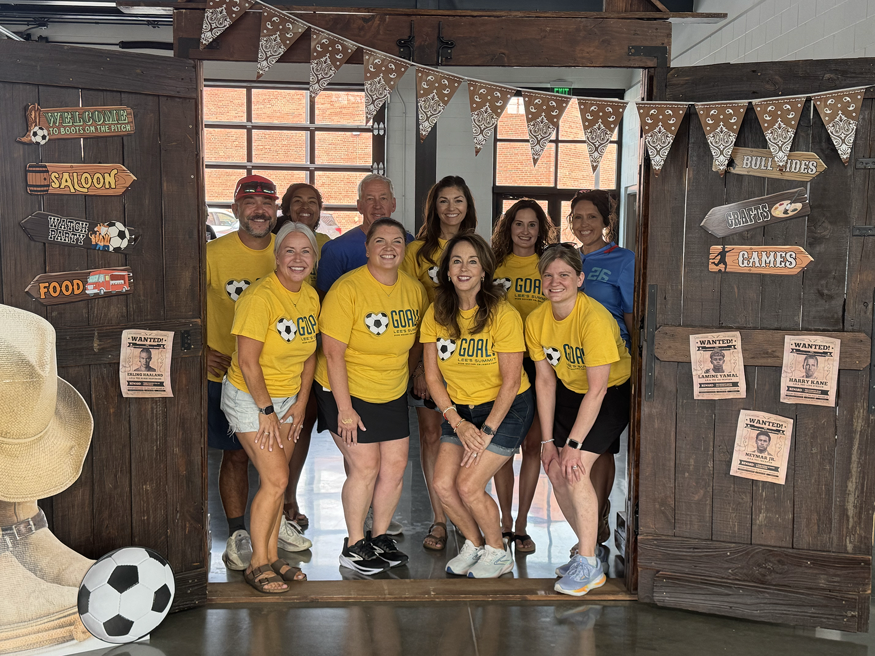 Group in yellow shirts posing in a rustic doorway with a soccer ball and festive bunting