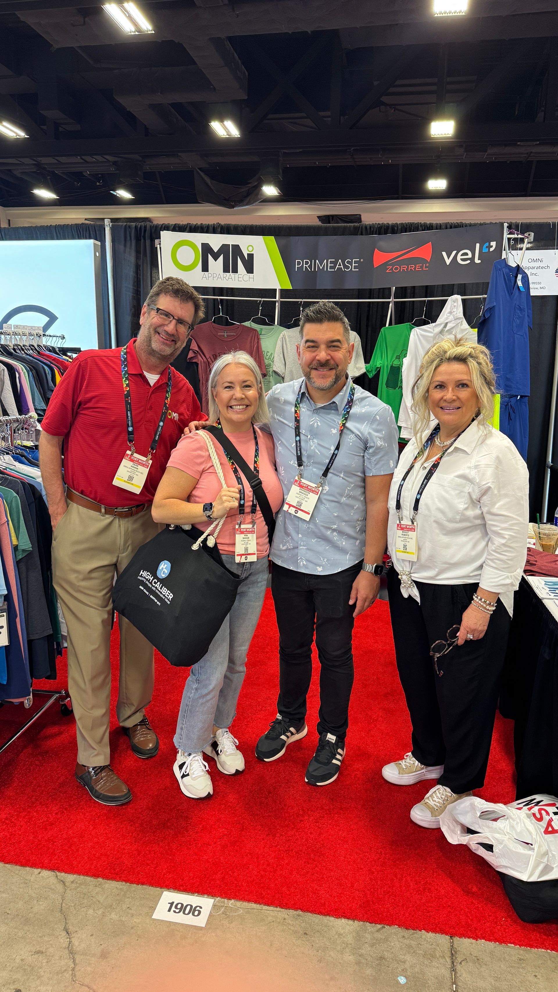 Four people smiling on a red carpet at a trade show booth, with banners and a black bag in front.