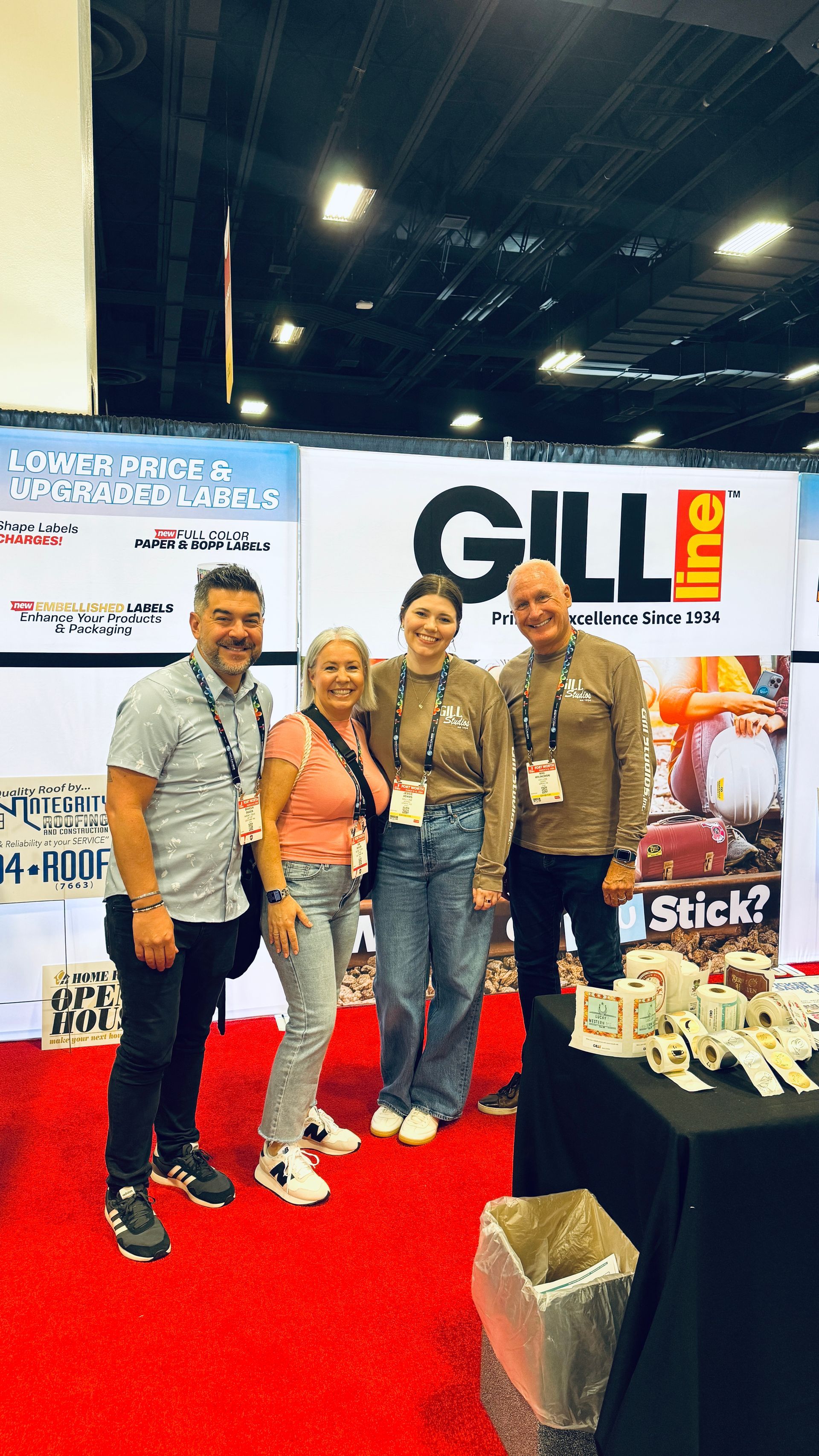 Four people stand at a trade show booth with a red carpet and “GILL” banner display.