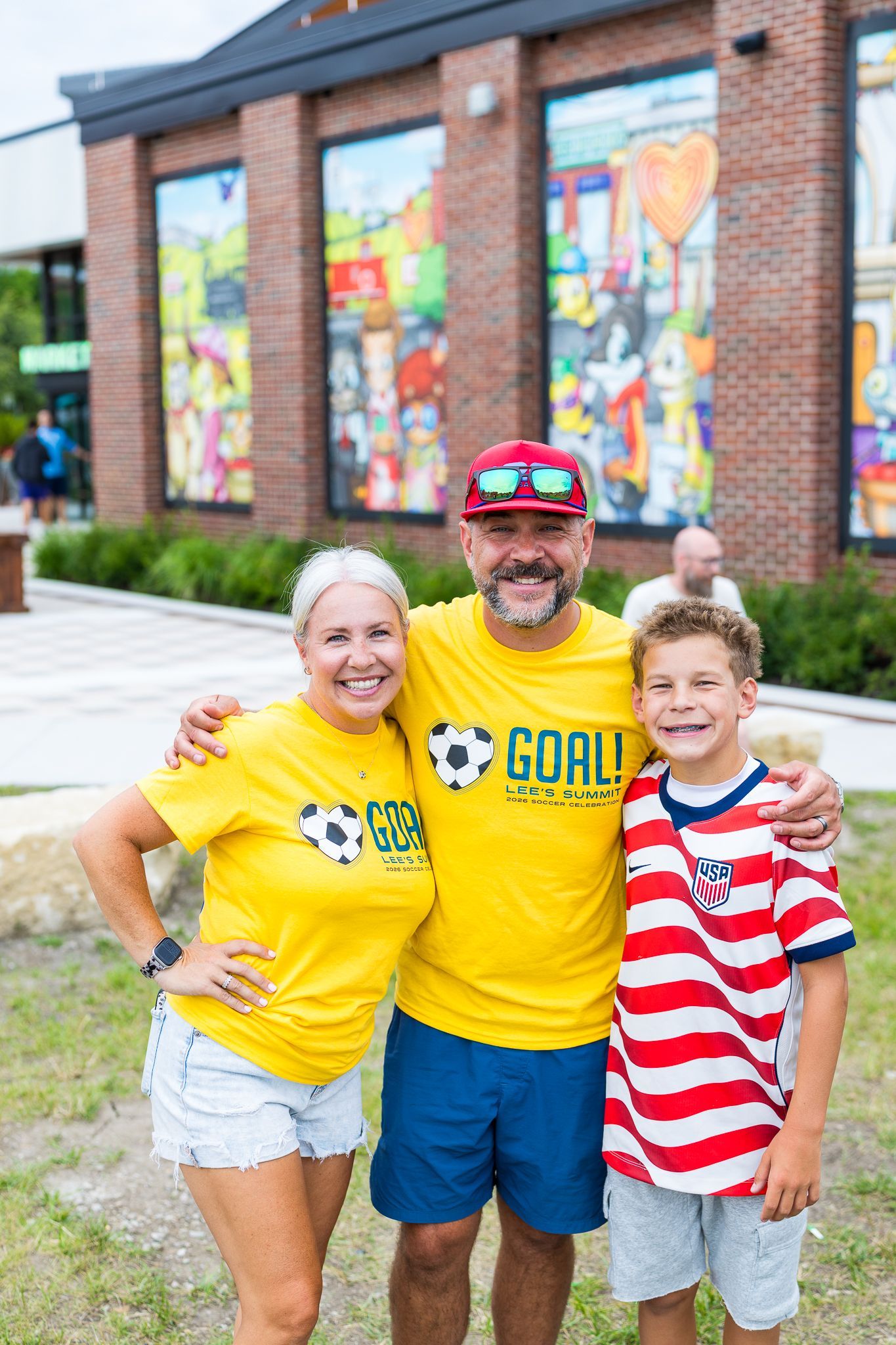 Three people in yellow shirts smiling and posing outdoors in front of a mural.