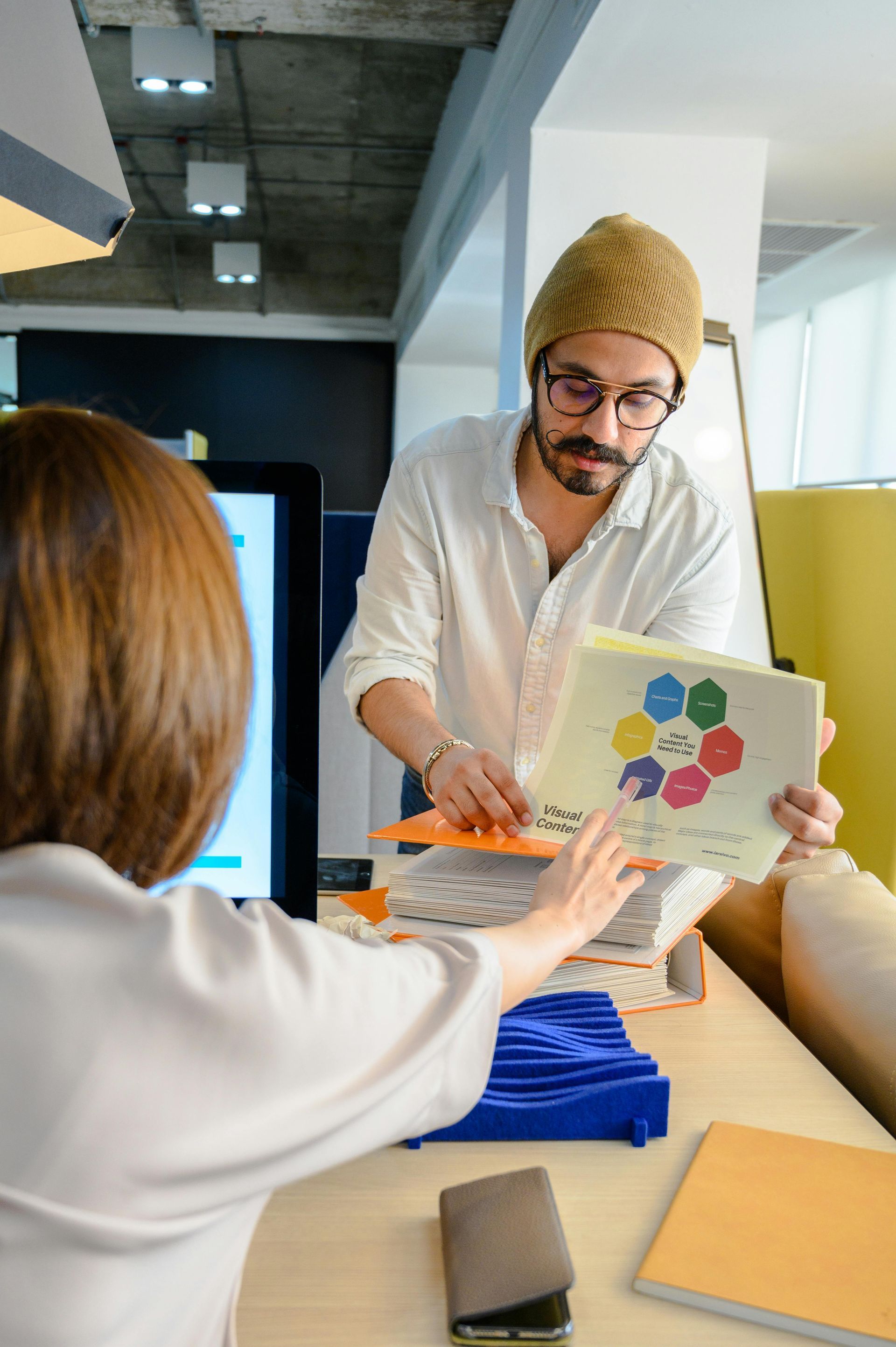Man in beanie hands a woman a chart in an office setting.