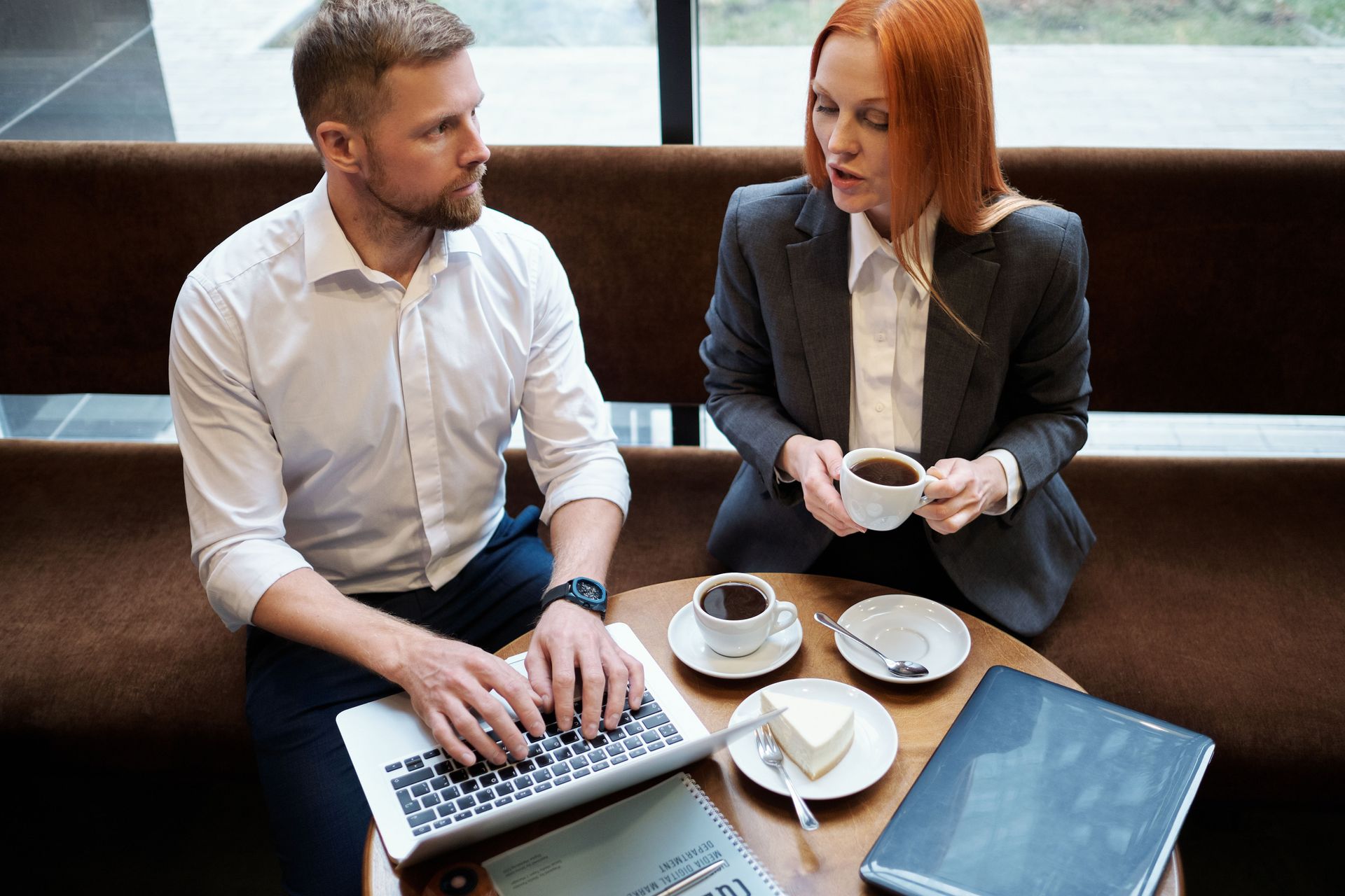 Two coworkers discuss over coffee at a table with a laptop and notebook.