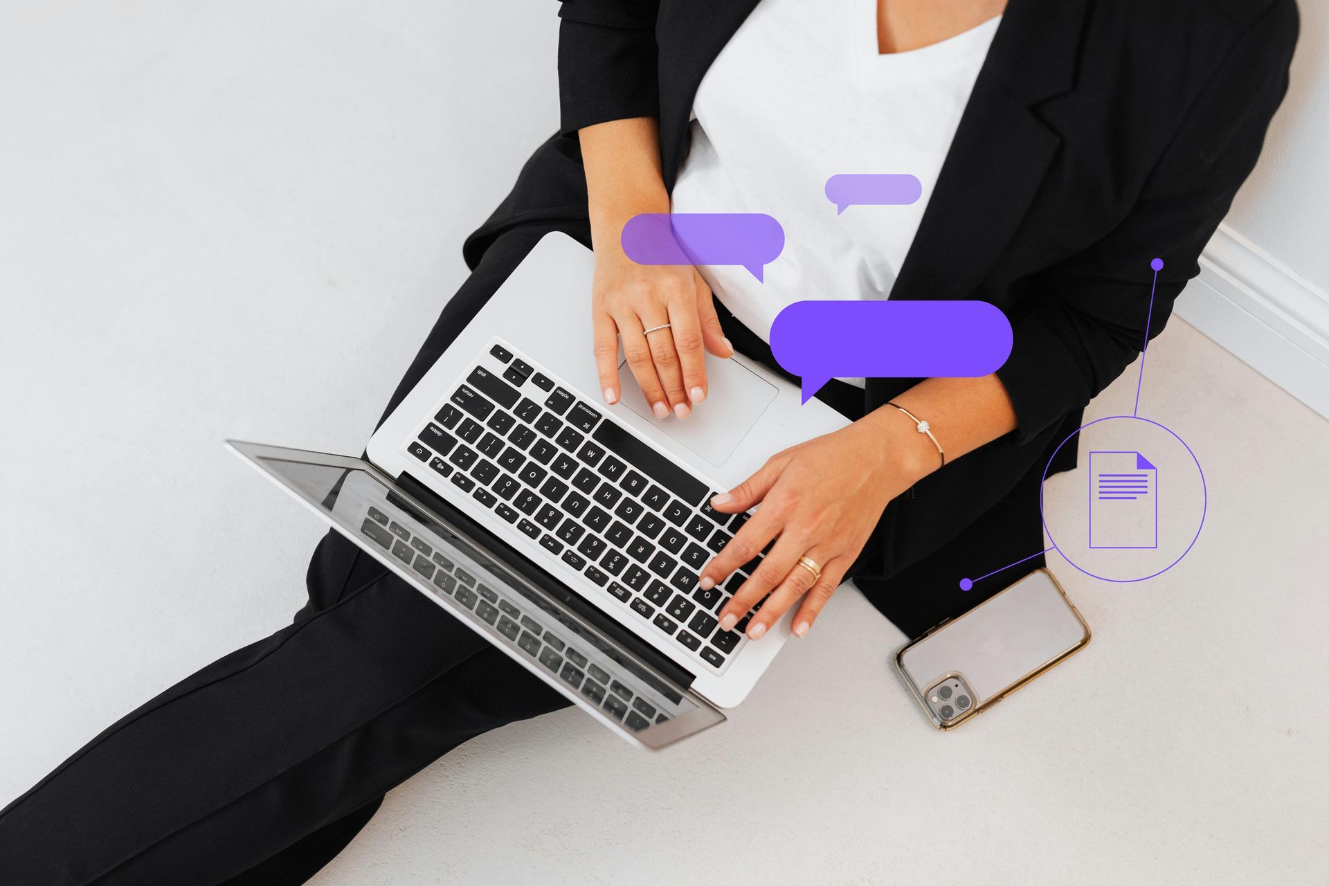 Woman in black suit uses laptop, chatting, sitting on floor. Phone nearby.