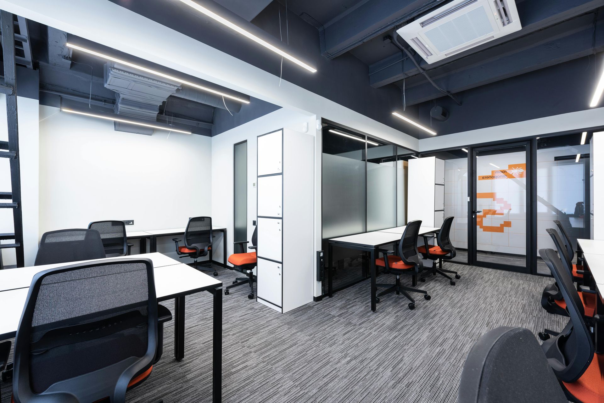 Modern office space with desks, chairs, and glass-walled rooms. Gray carpet, white desks, orange and black chairs.