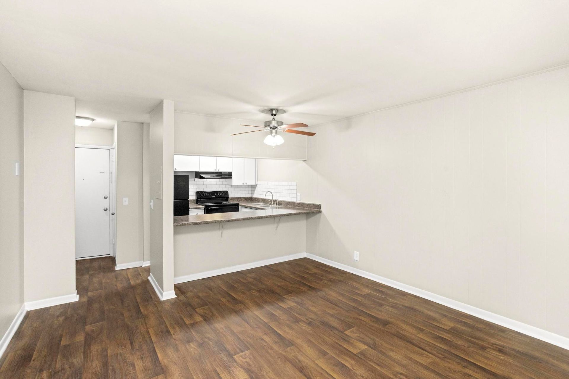 Empty apartment living area with dark wood floors and a small open kitchen.