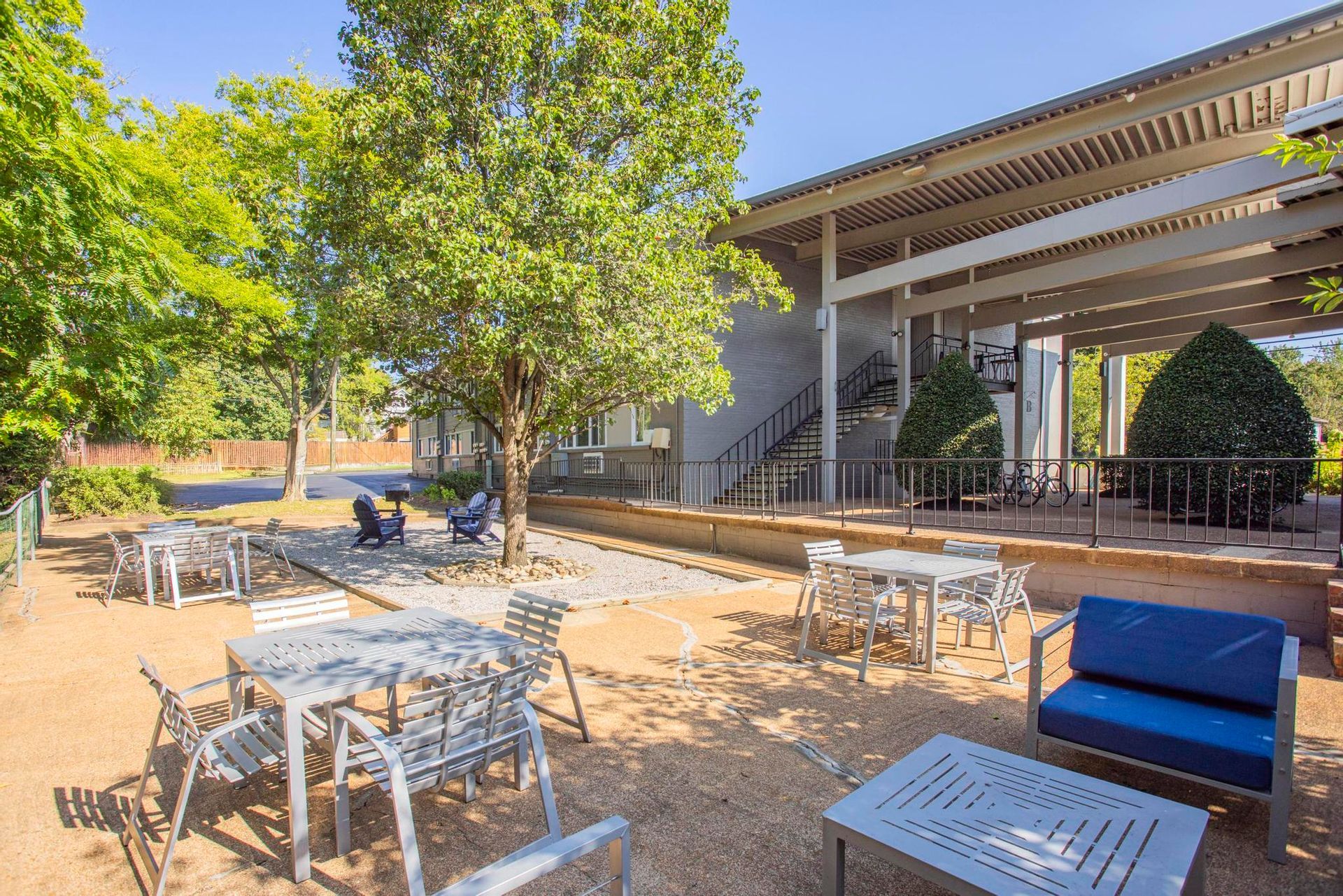 Outdoor patio with white tables and chairs beside a modern building, shaded by trees and an awning.