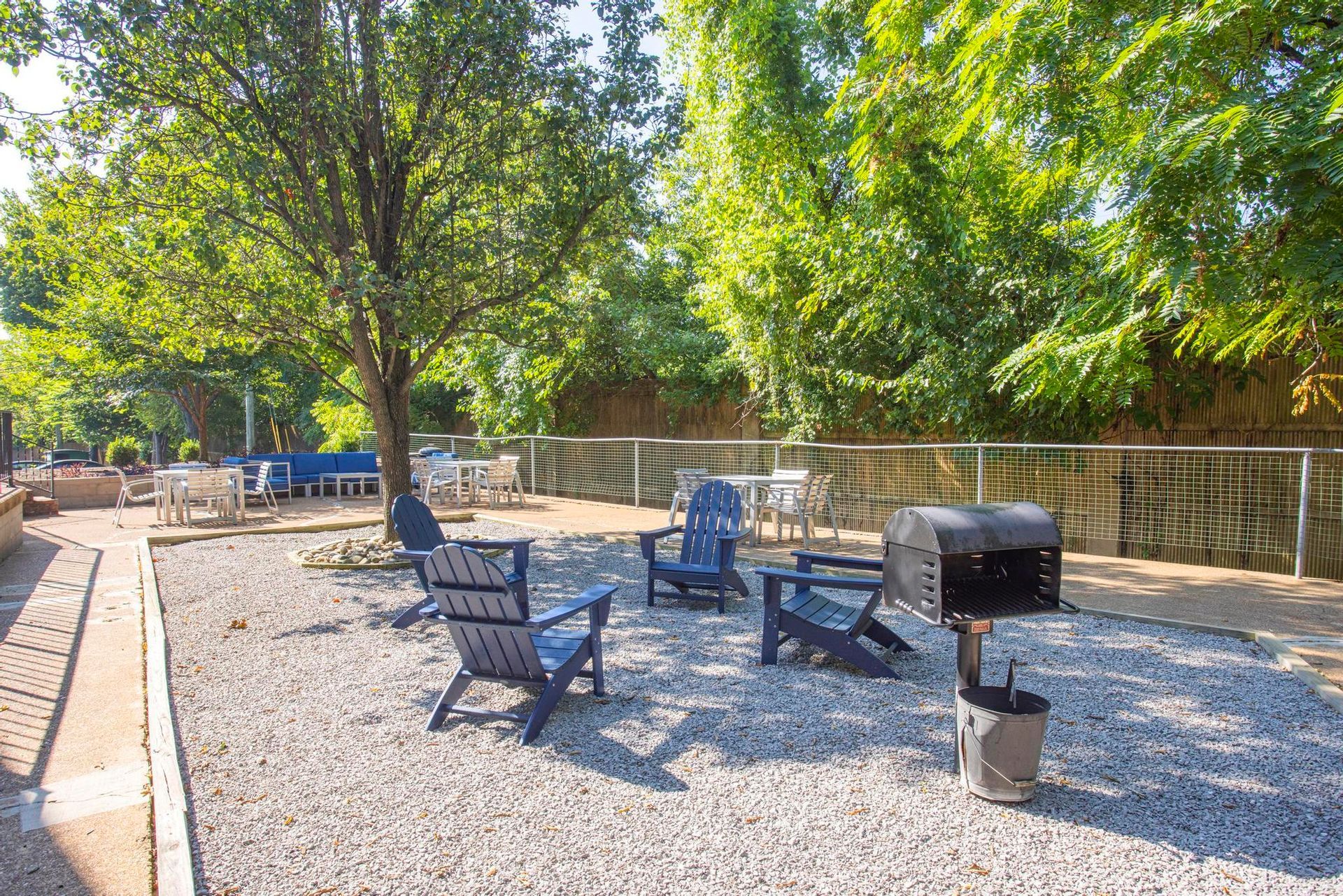 Gravel patio with chairs around a fire pit under trees and a wooden fence