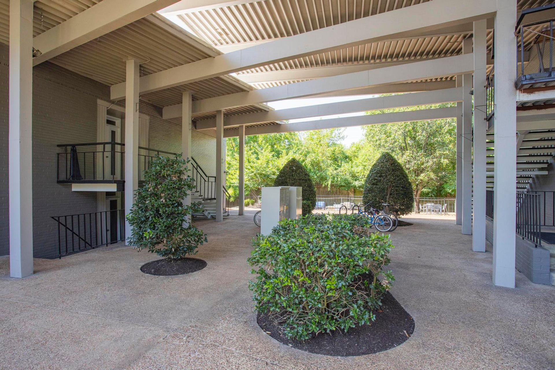 Covered outdoor walkway with white beams, potted shrubs, and a pebble floor in bright daylight