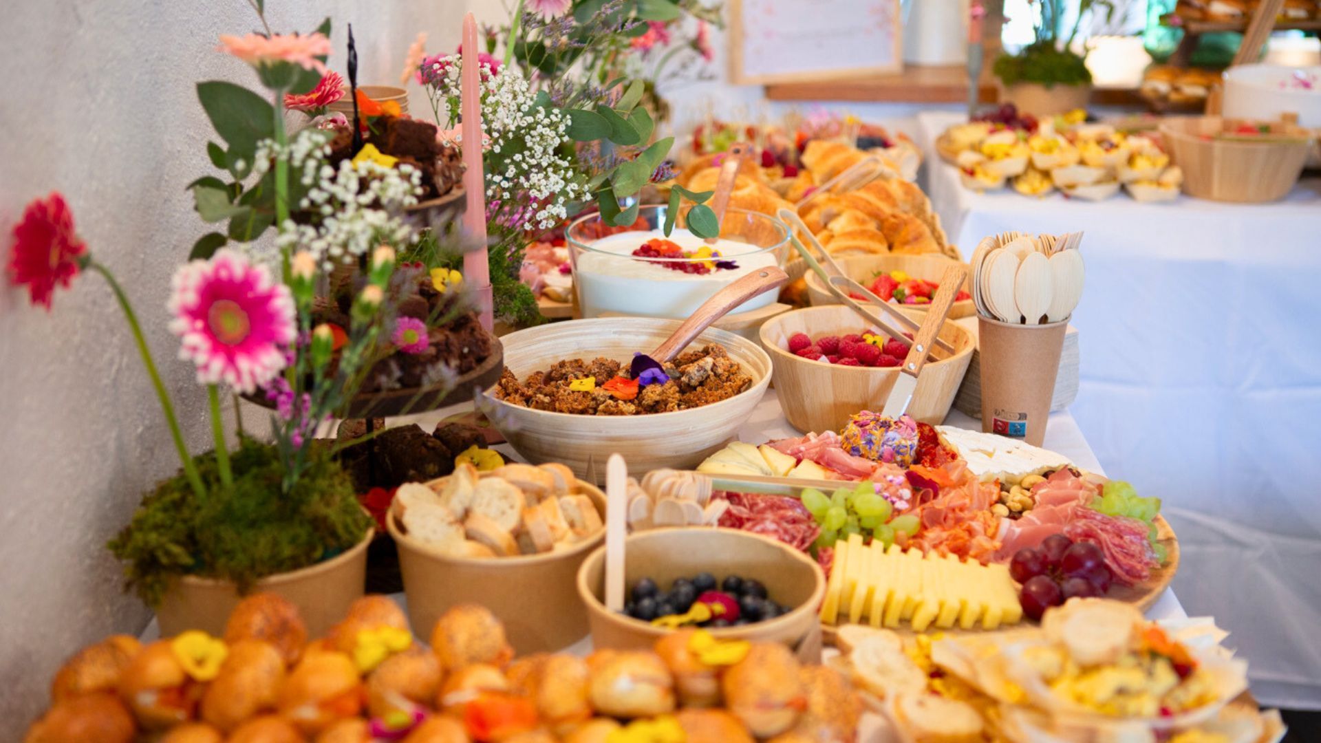 Un buffet de brunch coloré proposant des bols de granola, de fruits, de viennoiseries et de charcuterie, décoré de fleurs fraîches.