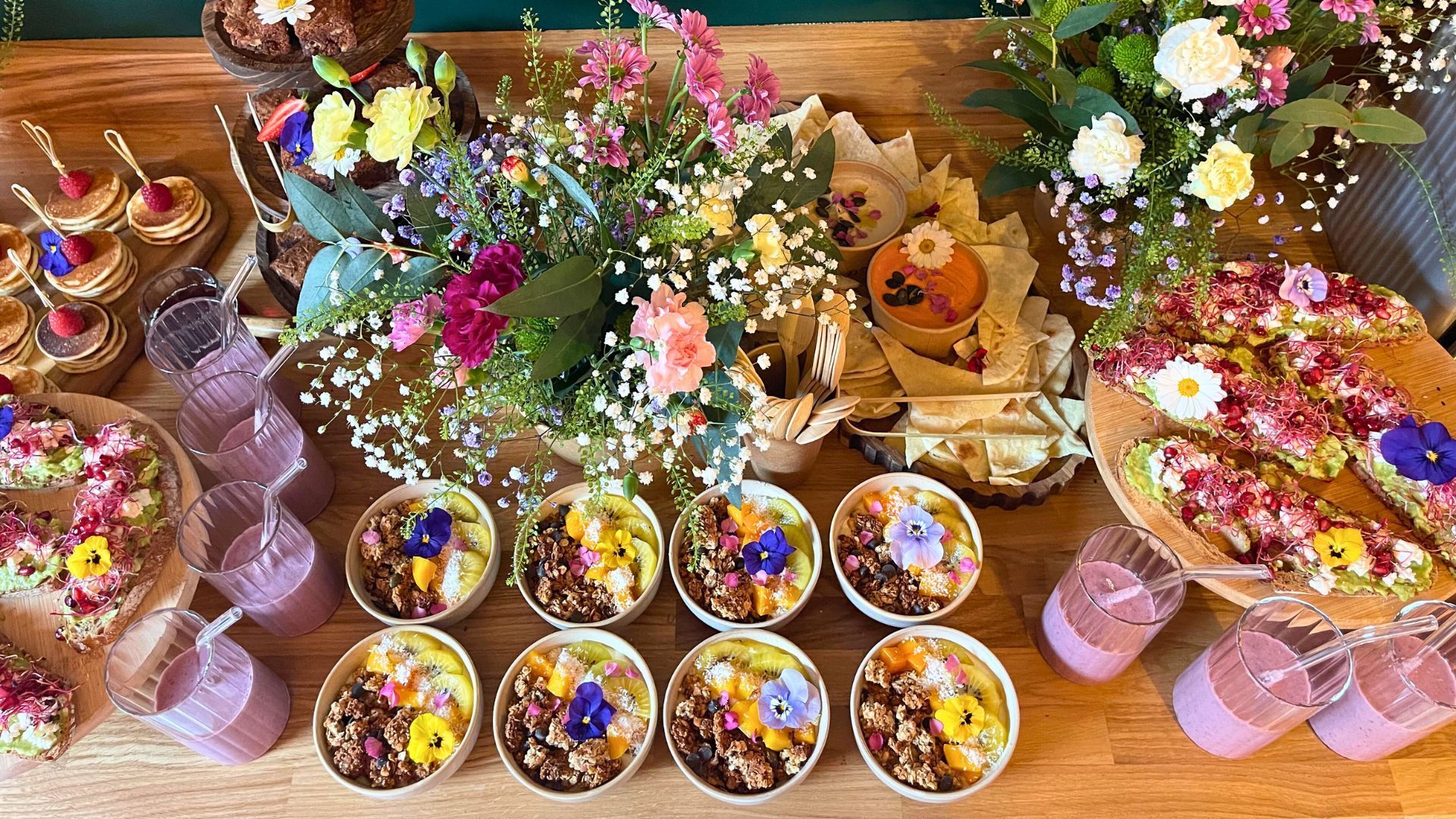 Une table en bois chargée de petits bols de yaourt garnis, d'amuse-gueules fleuris et de smoothies roses, le tout décoré de fleurs.