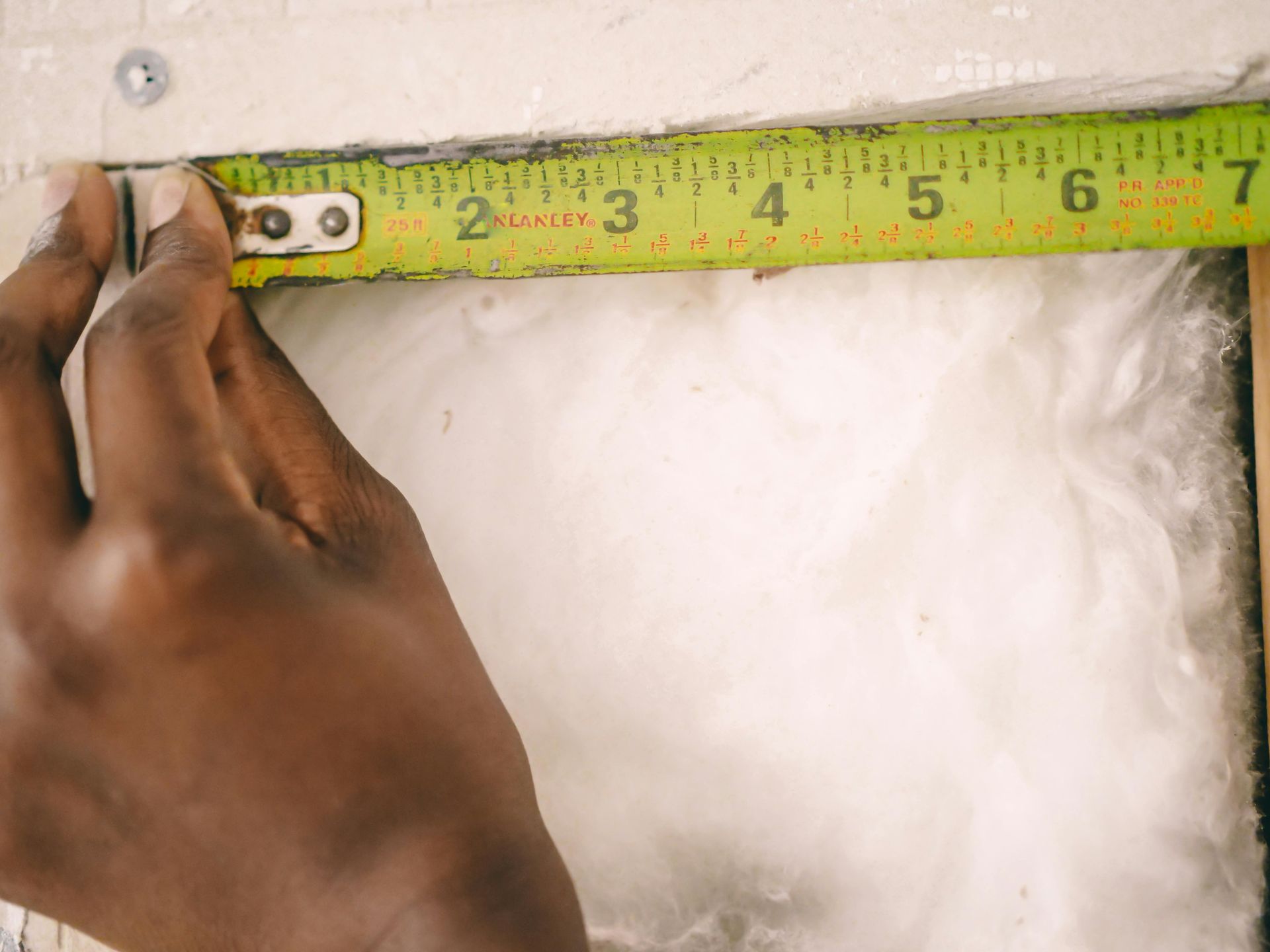 A hand measures an opening in a wall with a yellow tape measure. The opening shows insulation.
