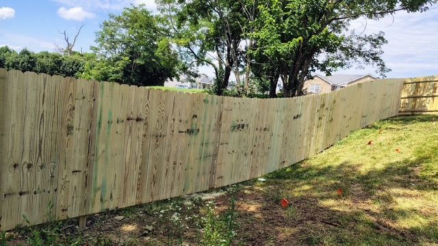 Wooden privacy fence curving along a grassy, tree-lined backyard slope under a bright sky