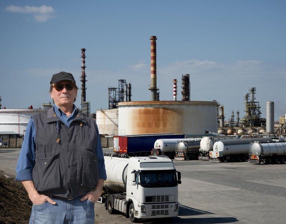A male driver standing in front of a Hazmat truck with a chemical plant on the background