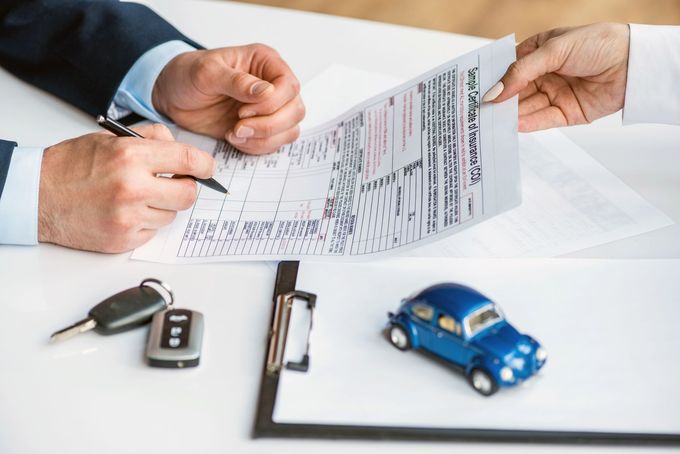 Una persona firma un documento di assicurazione auto mentre è seduta a una scrivania, con una macchinina blu giocattolo e le chiavi nelle vicinanze.