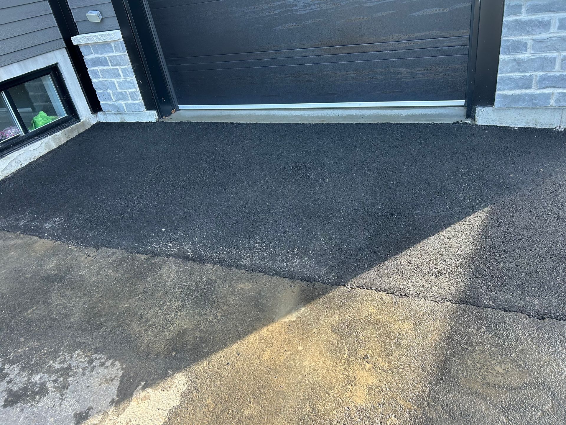 A newly paved section of black asphalt meeting older, weathered pavement in front of a house garage.