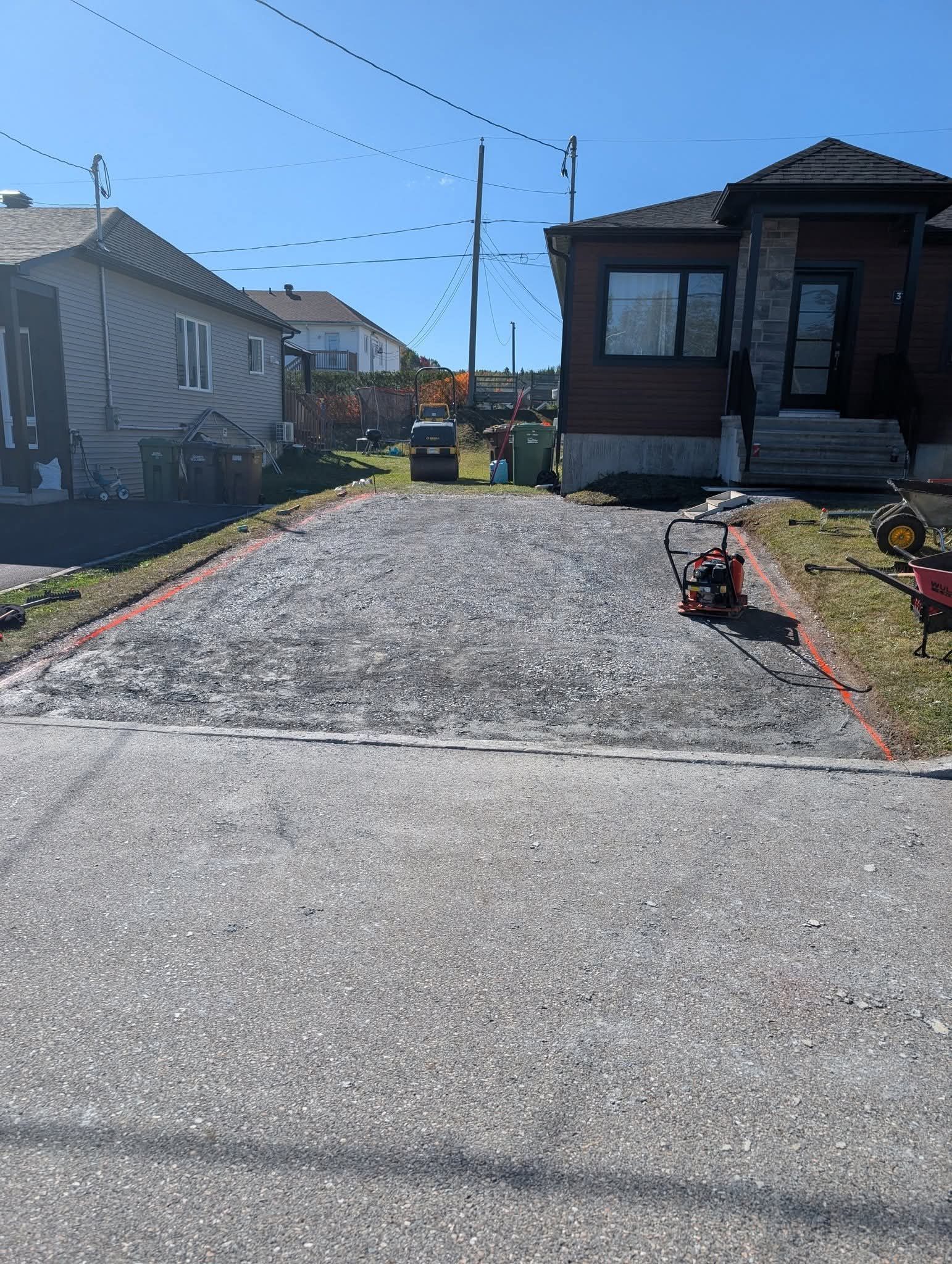 Une allée de gravier en construction, bordée de pierres rouges, entre deux maisons par une journée ensoleillée.