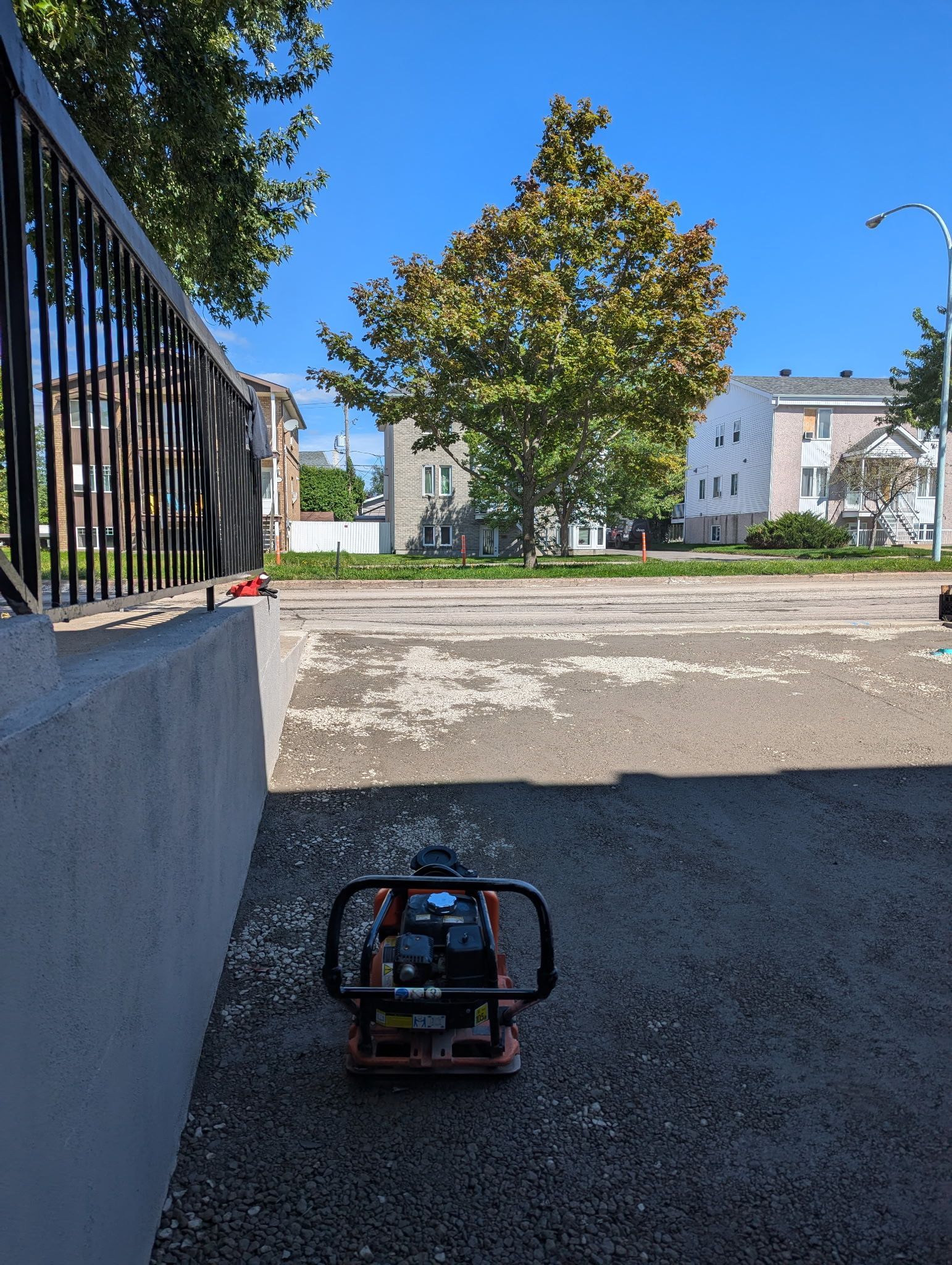 Une plaque vibrante est posée sur un terrain gravillonné, à côté d'un muret en béton entouré d'une clôture métallique noire, par une journée ensoleillée.