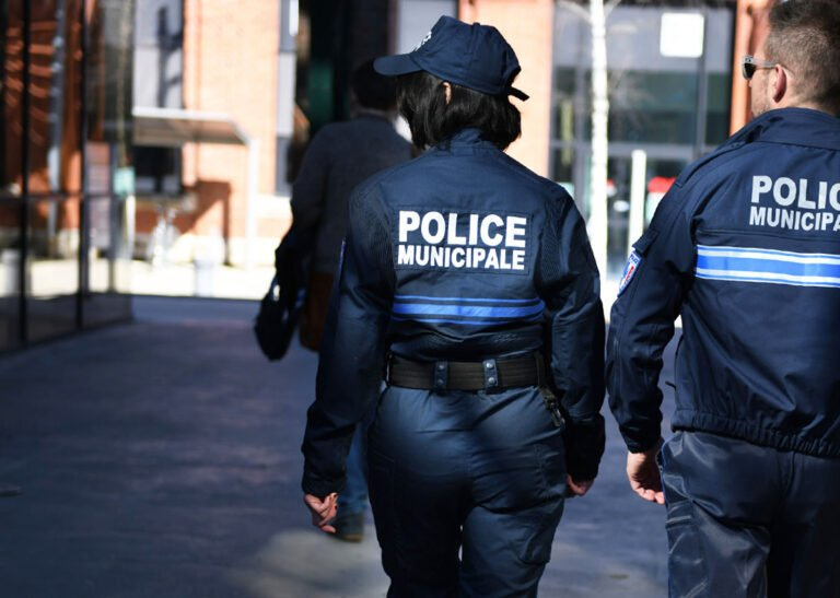 Deux agents de police municipale marchent à l'extérieur ; uniformes bleus avec l'inscription « POLICE MUNICIPALE » dans le dos.