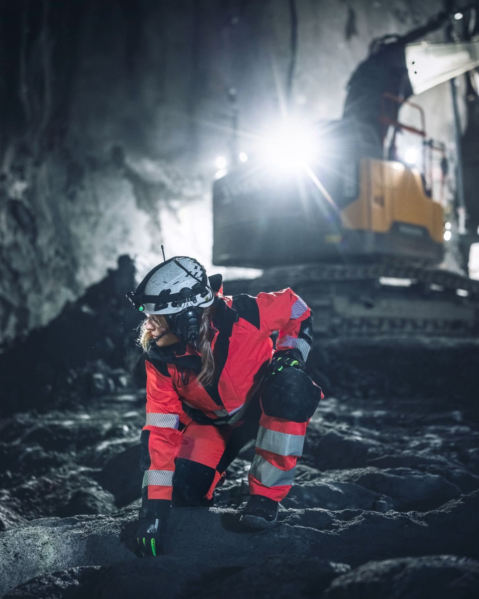 Une femme en combinaison de travail orange est agenouillée sur un sol sombre dans un tunnel éclairé par une lumière vive et bordé d'engins de chantier.