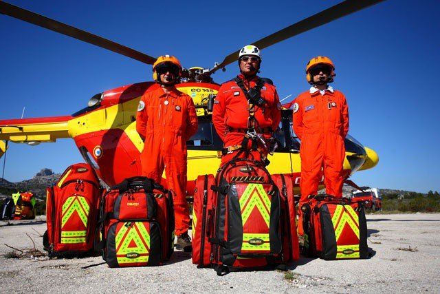 Trois secouristes en combinaisons orange se tiennent devant un hélicoptère, des sacs médicaux devant eux.