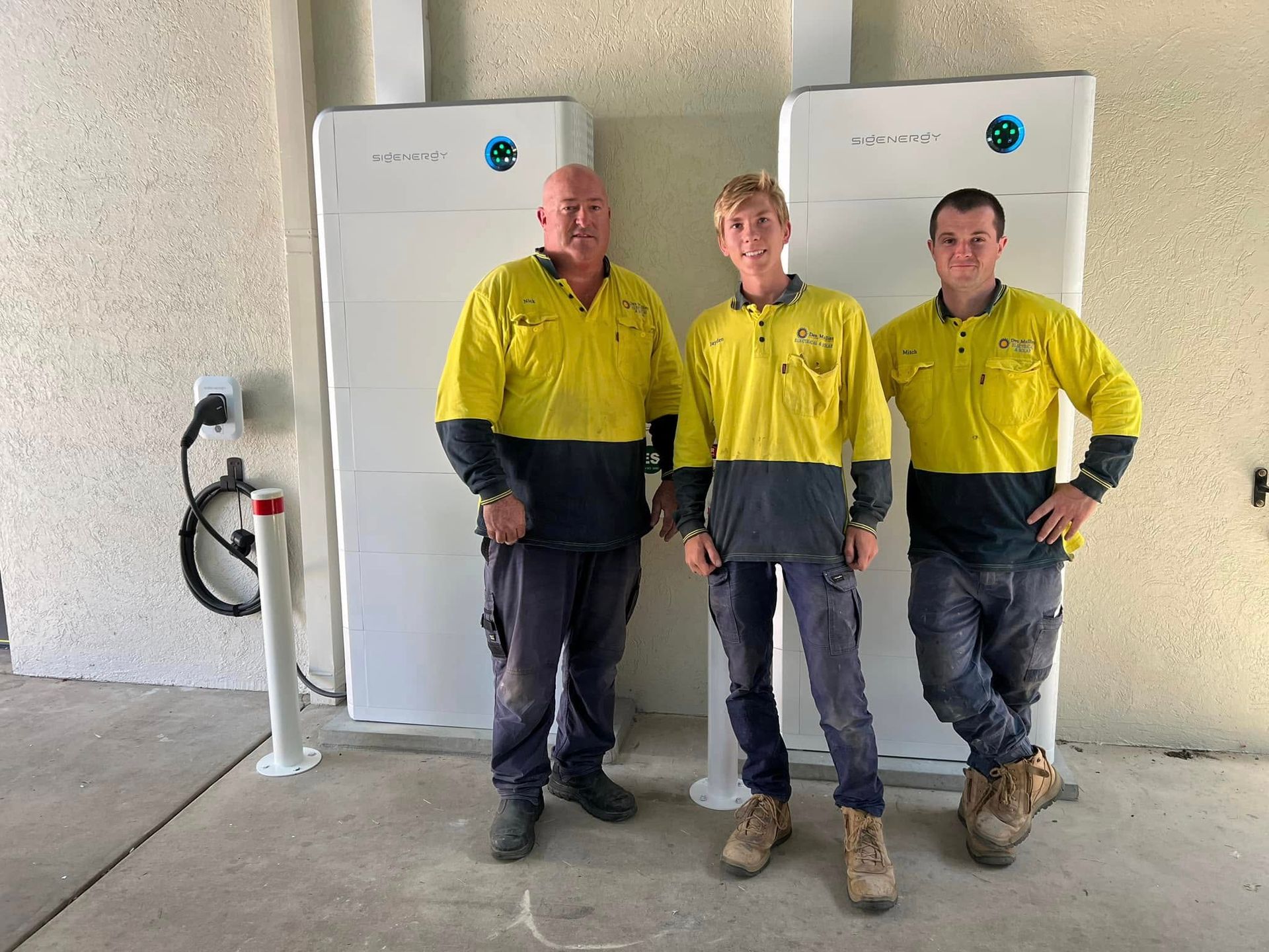 Three People in Work Uniforms Pose Near Solar Battery Storage Units — Des Mullins Electrical & Solar in Wagga Wagga, NSW