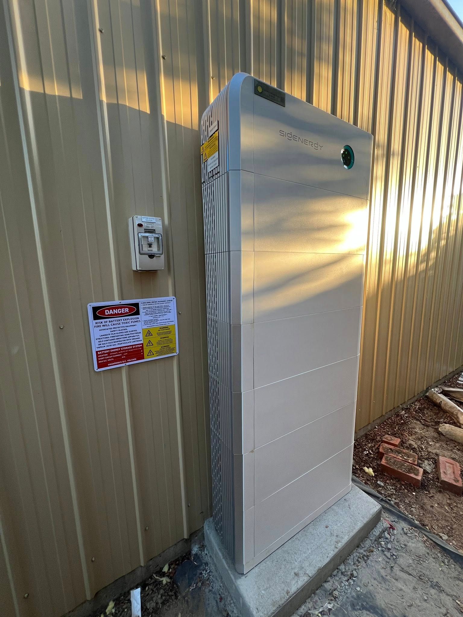 Metal Electrical Box on a Concrete Base Next to a Building Wall With a Warning Sign — Des Mullins Electrical & Solar in Wagga Wagga, NSW