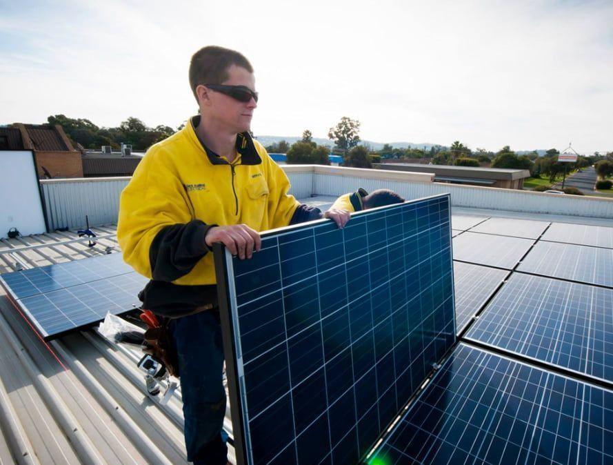 Man in Yellow Jacket Installs Solar Panel on a Rooftop — Des Mullins Electrical & Solar in Wagga Wagga, NSW