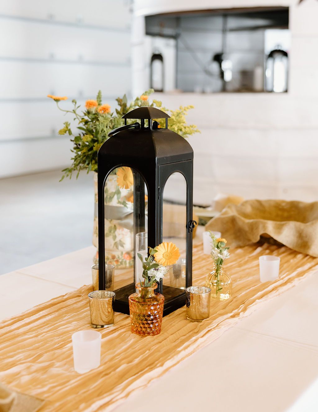 Black lantern centerpiece with flowers and candles on a white table runner