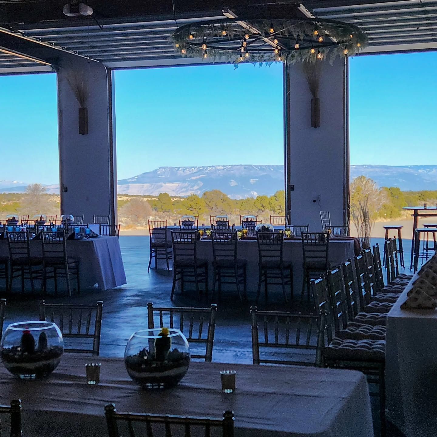 Open-air dining hall with tables and chairs overlooking mountains and a lake under a bright blue sky
