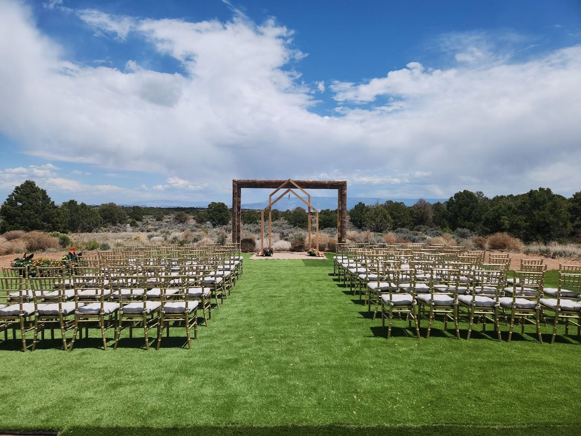 Outdoor wedding setup with rows of chairs lining a green aisle leading to a wooden arch under a blue sky