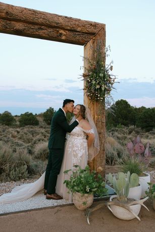 Bride and groom kissing beneath a rustic wooden arch in a desert landscape at sunset