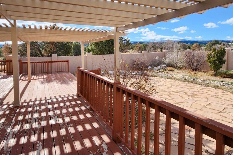 Sunny backyard deck with pergola, wooden railing, and stone patio under a blue sky