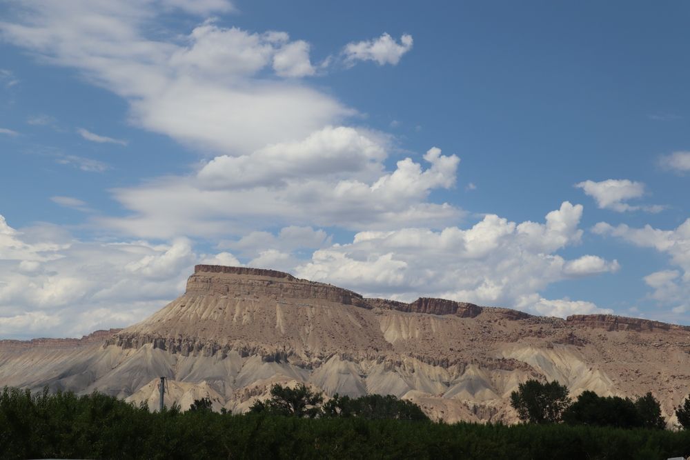 Stratified desert mesa beneath a partly cloudy blue sky, with trees in the foreground