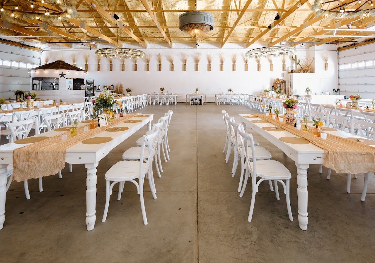 Brightly decorated banquet hall with long white tables, chairs, and floral centerpieces in a rustic setting
