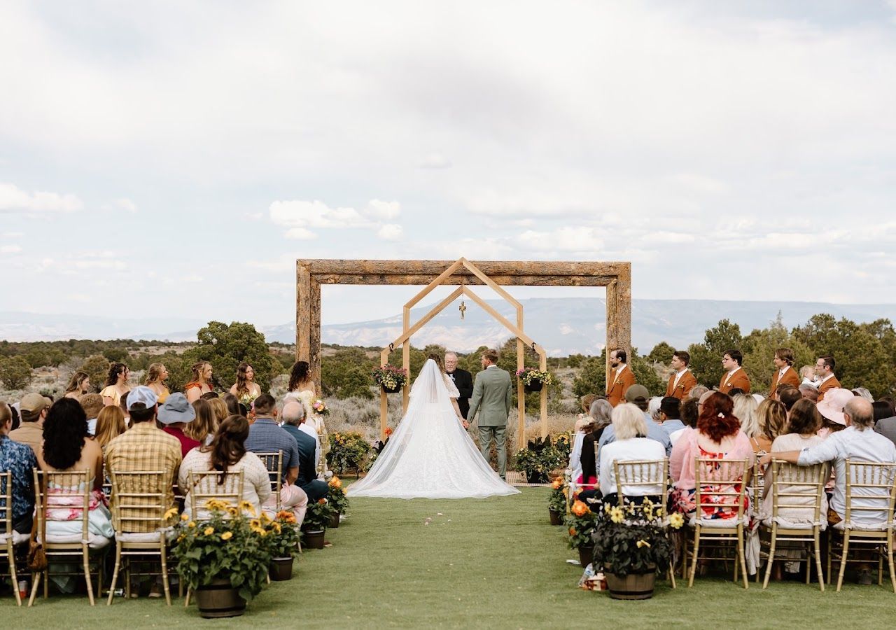 Bride and groom at a rustic outdoor wedding ceremony beneath a wooden arch, guests seated on either side.
