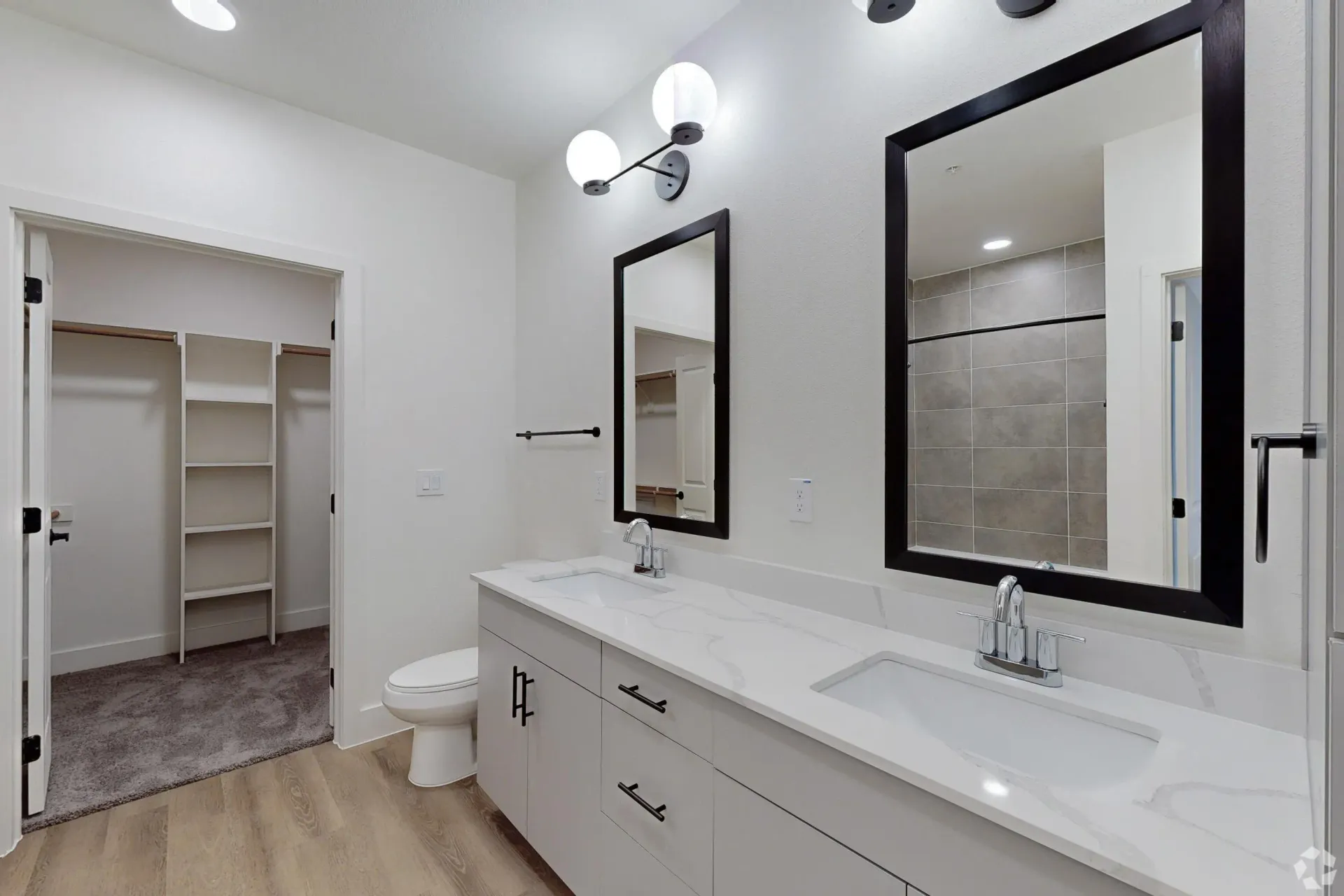 Double vanity with white quartz countertop, modern light fixtures, and large mirrors above. A walk-in closet is visible to the left.