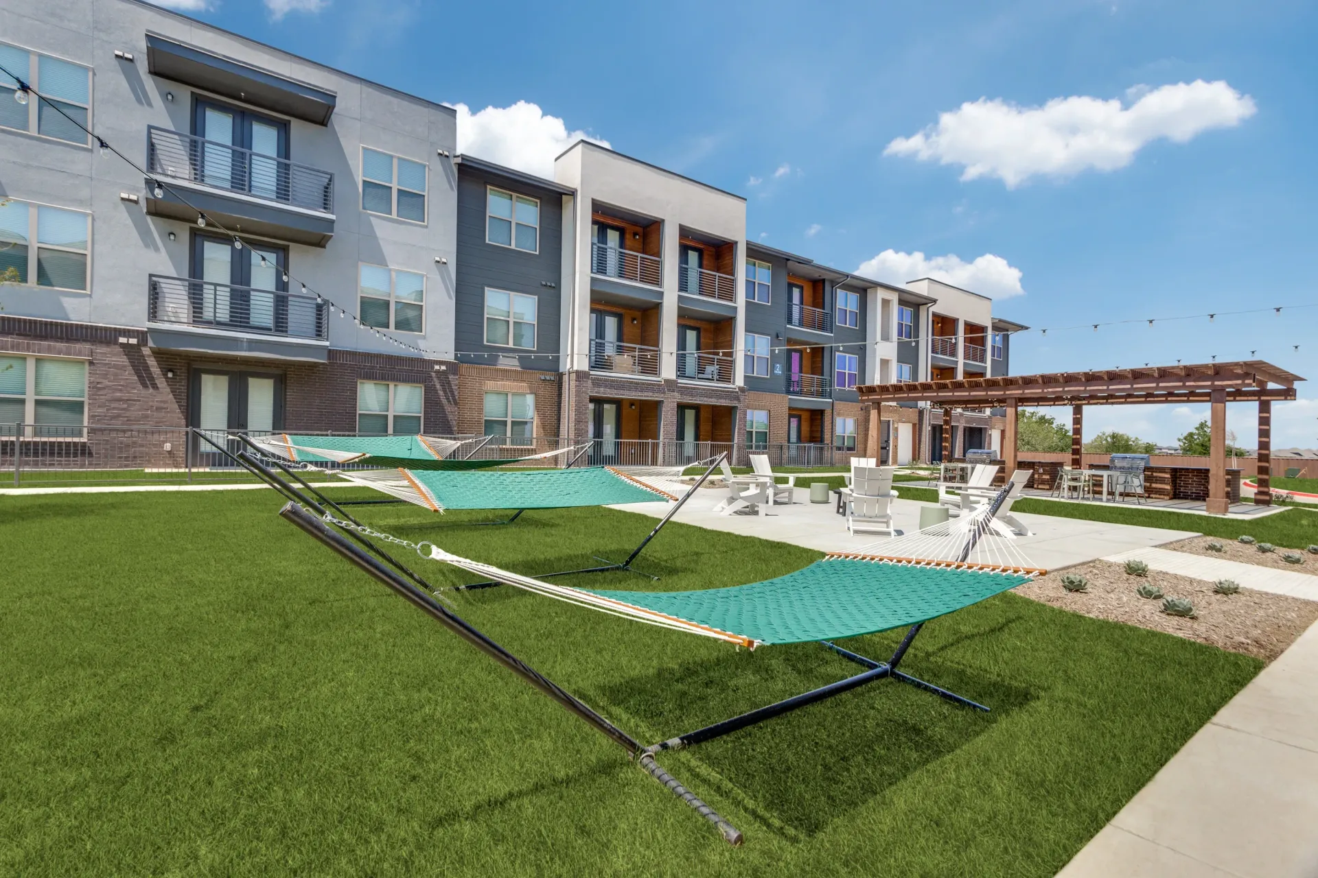 Outdoor area with hammocks, lounge chairs, and a grilling station next to a modern apartment building.