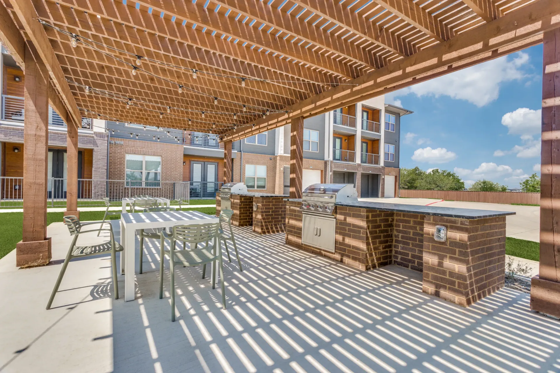 Outdoor grilling area with built-in grills, tables, and chairs under a wooden pergola with string lights.