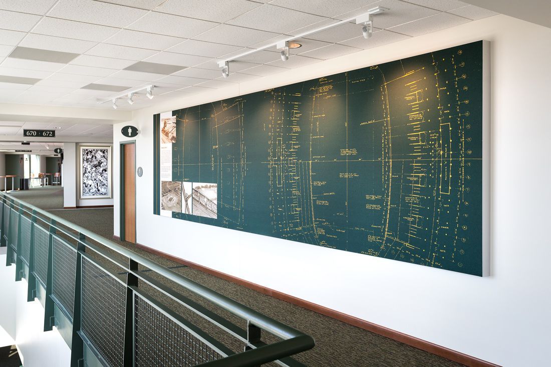 Hallway with artwork of a city skyline lit up. Metal railings on the left, fluorescent lighting, and white walls.