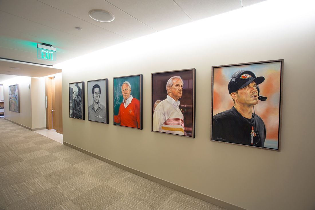 Hallway with five portraits of people on the wall; light beige walls and carpet, fluorescent lighting.