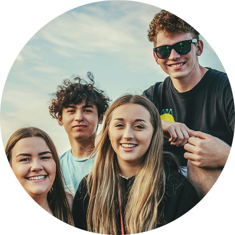 Four smiling friends pose closely together outdoors under a bright sky.