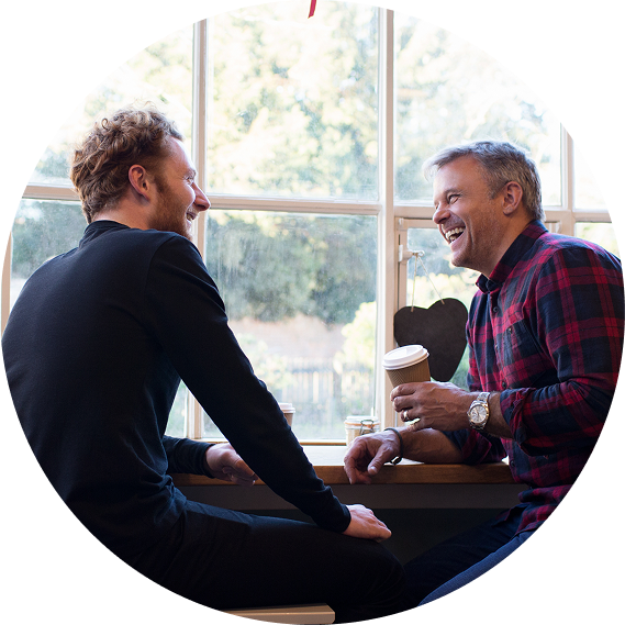 Two people chatting by a bright window, one holding a coffee cup and smiling.