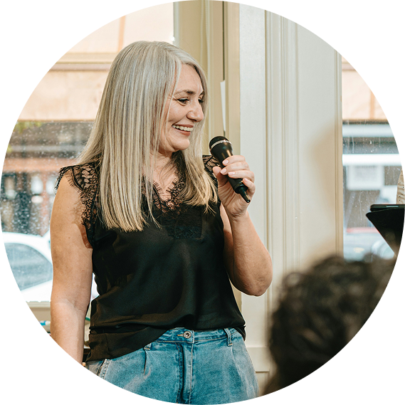 Woman speaking into a microphone at an indoor event, wearing a black top and jeans, smiling.