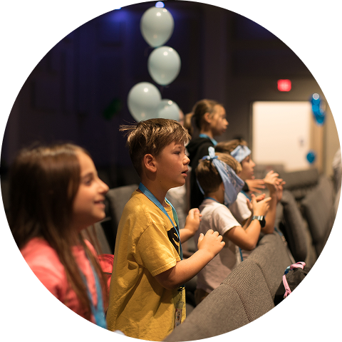 Children standing and clapping in a dim auditorium, with blue balloons and a stage in the background