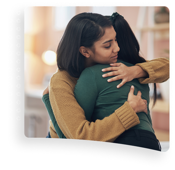Two people hugging warmly indoors, one in a green sweater and one in a tan sweater.