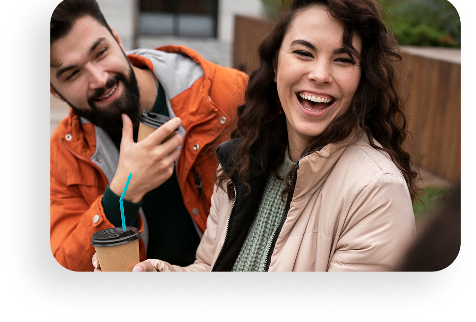 Smiling couple outdoors, one holding a drink and wearing orange, the other in a beige coat and scarf