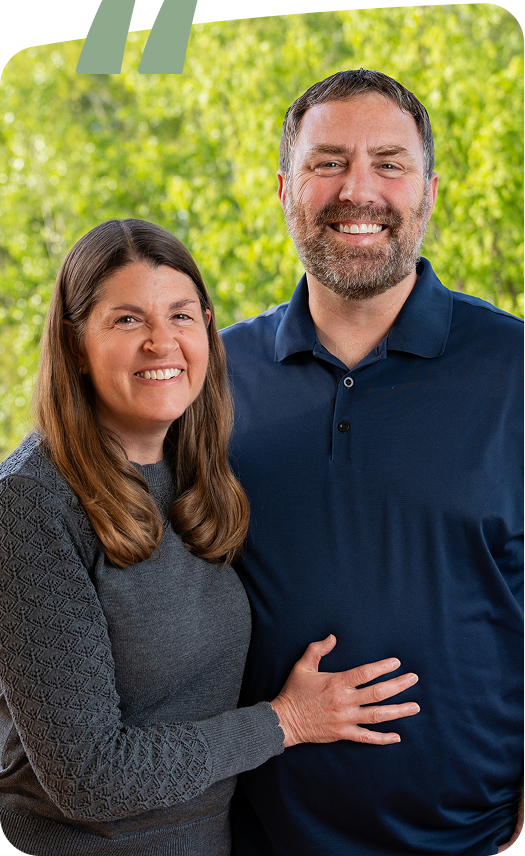 Smiling couple standing outdoors, with trees blurred in the background.