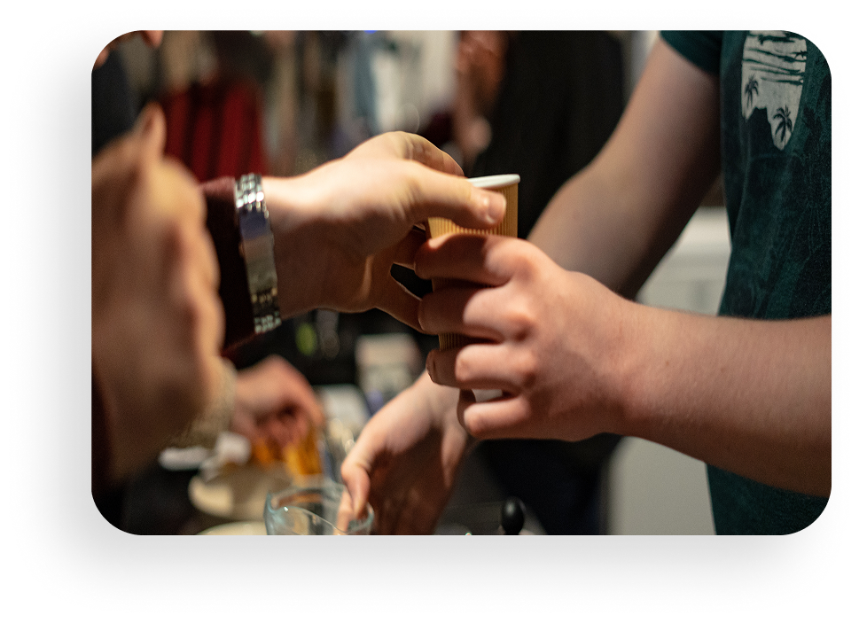 Hands passing a drink at a social gathering, with people reaching in from both sides.