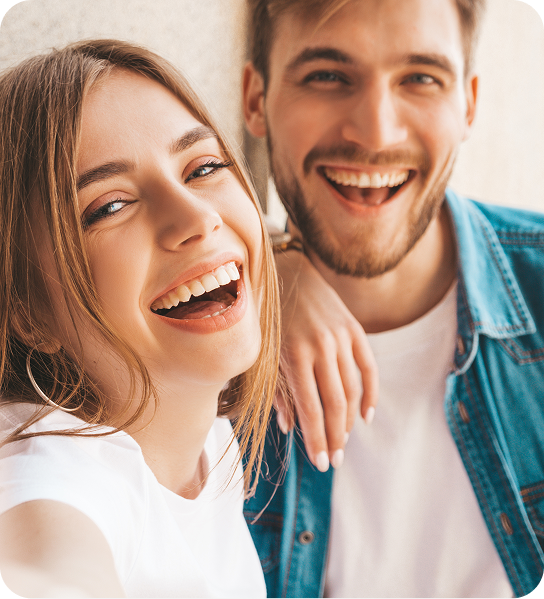 Two smiling friends posing close together indoors, one in a white shirt and the other in a denim jacket.